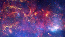 A multiwavelength view of the Milky Way center displaying blue, purple, yellow, and orange colors, which show gas and dust of various shapes and textures, with cliff-like ridges and stars of different colors spread throughout.