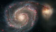 A visible wavelength view of the Whirlpool Galaxy, which shows a spiral galaxy filled with a bright yellow core of gas and dust and brown dusty arms with pink and blue stars within, and a smaller yellow galaxy to the top right.