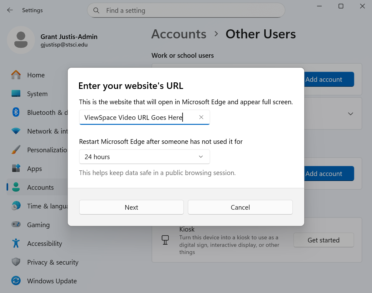 Windows 11 Settings window under "Accounts" > "Other Users." A dialog box titled "Enter your website's URL" is open, prompting the user to input a URL that will launch in Microsoft Edge in full-screen kiosk mode. The placeholder text in the input field reads "ViewSpace Video URL Goes Here." Below, there's a dropdown menu labeled "Restart Microsoft Edge after inactivity," set to “24 hours." Two buttons "Next" and "Cancel" are at the bottom of the dialog.